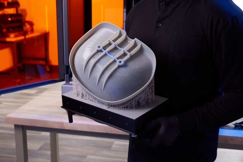 黒い手袋をした手が、レジンで濡れた大きなグレーの造形品を乗せたビルドプラットフォームを持っている様子