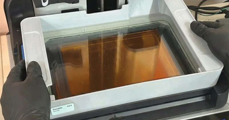 手袋をした両手で、レジンの入ったタンクを Form 4 から水平に引き出している様子