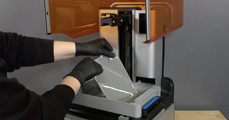 黒い手袋をした両手が、プリンター内のタンク底面から透明な硬化シートをめくり上げている様子